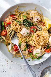 GVL A plate of Chicken Scampi spaghetti with chunks of chicken, cherry tomatoes, spinach, fresh herbs, and a lemon wedge, with a fork twirled in the noodles on a light background.