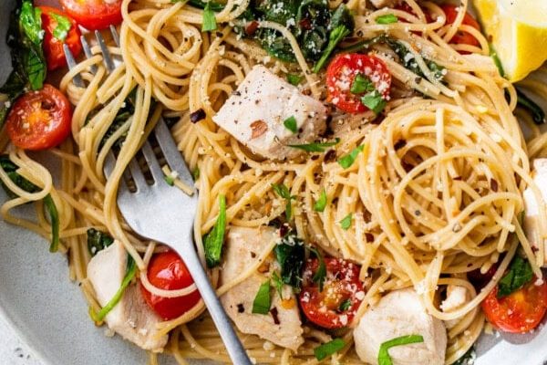GVL A plate of Chicken Scampi spaghetti with chunks of chicken, cherry tomatoes, spinach, fresh herbs, and a lemon wedge, with a fork twirled in the noodles on a light background.