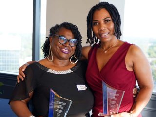 GVL Two women, proud award winners, smile and pose together indoors, each holding a clear glass award. One wears glasses and a black top; the other a sleeveless burgundy dress. They stand before a city-view window at the Empowering Entrepreneurship event.