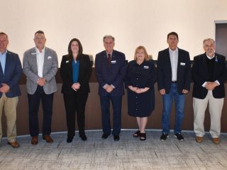 GVL Seven people stand in a row inside a building, dressed in business or business casual attire, posing for a City Council group photo against a neutral-colored wall, highlighting unity on the Simpsonville budget and commitment to no tax increase.