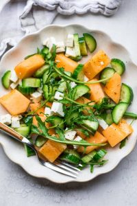 GVL A refreshing salad featuring sweet melon and crisp cucumber slices, tossed with arugula and feta cheese cubes, seasoned with pepper, and served with a fork on a white plate.
