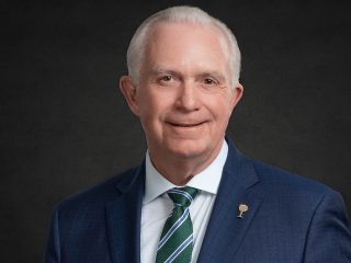 GVL An older man with short white hair, wearing a dark blue suit, white shirt, and green striped tie, smiles in front of a dark background—embodying the lasting legacy of President Miller at Greenville Tech.