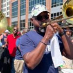 GVL A man wearing sunglasses and a hat plays the trombone in an outdoor brass band performance at the Greenville Jazz Fest, with other musicians and a city building visible in the background.