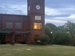 GVL A brick building with a clock tower is shown at dusk. The clock reads 7:20, and some windows are warmly lit as the City Council discusses the Greer budget. Trees and a grassy lawn surround the building under a partly cloudy sky.