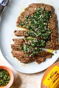 GVL Sliced grilled chimichurri steak topped with fresh sauce on a white platter, served alongside corn on the cob and a small bowl of extra chimichurri sauce.