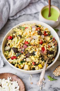 GVL A bowl of Mediterranean Couscous Salad with grilled vegetables like red peppers and zucchini, served with a fork. A small pitcher of green dressing and a plate of crumbled feta cheese sit nearby on a marble surface.