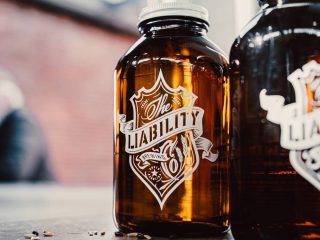 GVL A close-up of a brown glass bottle with a decorative label that reads The Liability in white script, crafted by Liability Brewing in Greenville. Another similar bottle is partially visible beside it.