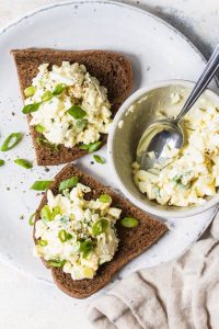 GVL Two slices of dark bread topped with creamy high protein egg salad and chopped green onions on a white plate, next to a bowl of lower fat egg salad with a spoon.