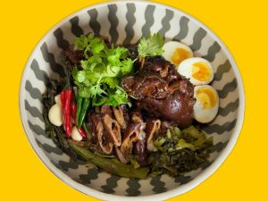 GVL A bowl of braised pork leg with boiled eggs, pickled greens, fresh cilantro, red and green chilies, and garlic, served in a patterned bowl on a yellow background—just like dishes at the best restaurants in Carroll Gardens and Cobble Hill.