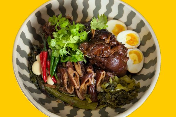 GVL A bowl of braised pork leg with boiled eggs, pickled greens, fresh cilantro, red and green chilies, and garlic, served in a patterned bowl on a yellow background—just like dishes at the best restaurants in Carroll Gardens and Cobble Hill.