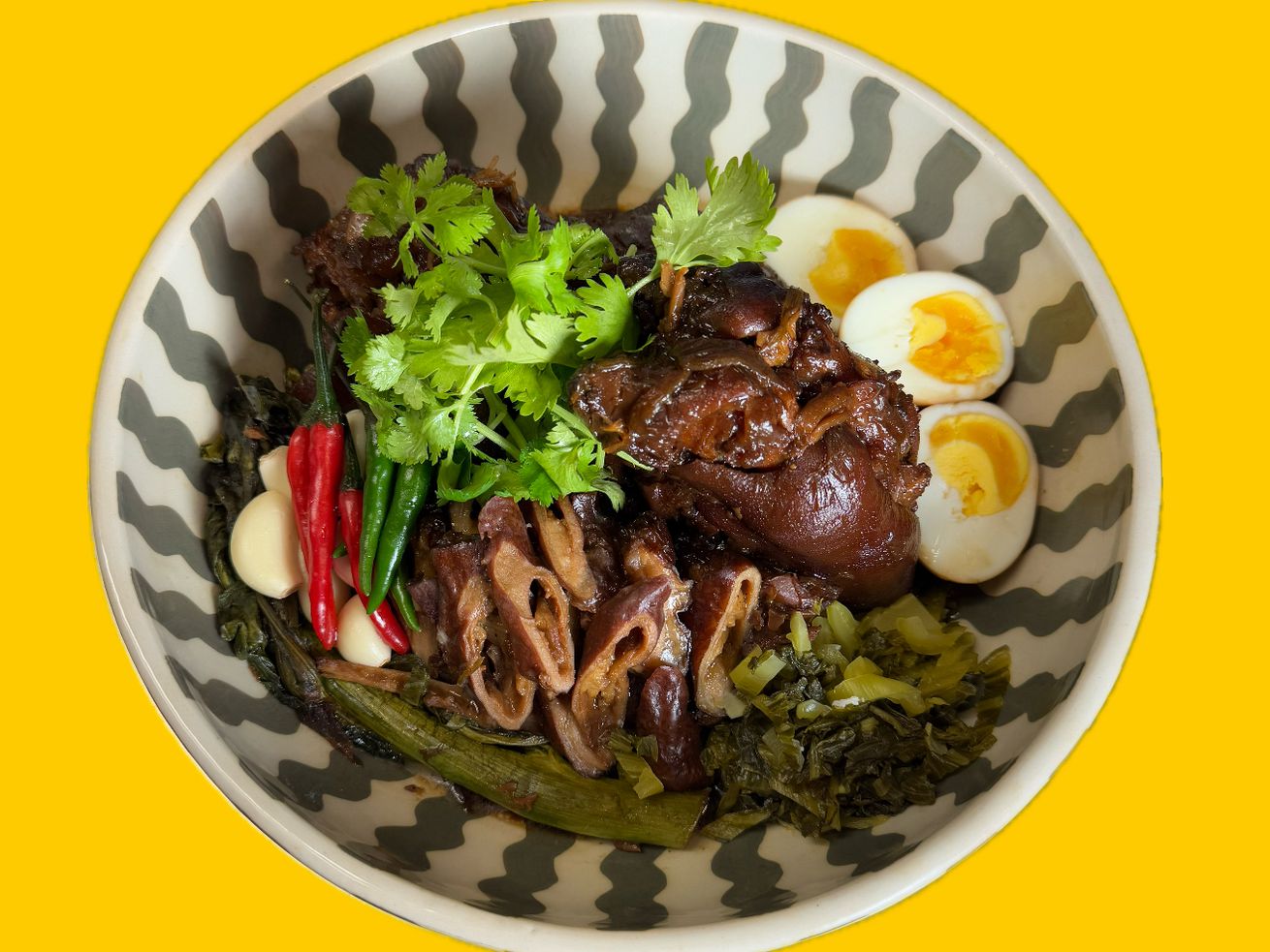 GVL A bowl of braised pork leg with boiled eggs, pickled greens, fresh cilantro, red and green chilies, and garlic, served in a patterned bowl on a yellow background—just like dishes at the best restaurants in Carroll Gardens and Cobble Hill.