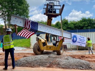 GVL Construction workers guide a steel beam, decorated with an American flag and a banner, during a topping-off ceremony at a construction site with trees and containers in the background.
