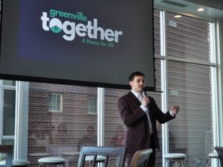 GVL A man in a suit speaks into a microphone in front of a screen displaying the Greenville Together: A Home for All logo, leading a community conversation on homelessness in a modern room with large windows and brick walls.