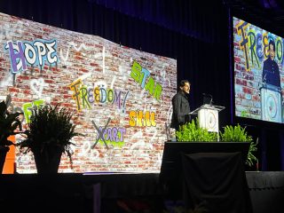 GVL A speaker stands at a podium on stage during the Family Effect annual breakfast, with a brick wall backdrop featuring colorful graffiti words like HOPE, FREEDOM, START SMALL, and EXTRA. The Phoenix Center event scene is shown on a large screen nearby.
