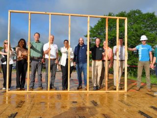 GVL Nine people stand side by side, smiling behind a wooden wall frame at a Habitat Greenville CEO Build construction site. With trees and a cloudy sky in the background, the wet ground and one hard hat hint at teamwork and progress.