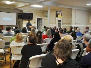 GVL A man in a suit speaks at a podium to a seated audience in a Greenville community hall. A screen beside him displays the word together as he addresses topics like homelessness and the areas housing surge. The audience listens attentively.