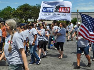 GVL A group of people in gray shirts participate in a Carry The Load memorial walk, carrying an American flag and a banner that reads, Carry The Load. Who are you carrying? Trees and buildings are visible in the background, honoring fallen service members.