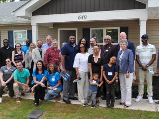 GVL A diverse group of adults and children pose and smile together in front of a beige Habitat for Humanity home with the number 640 above the entrance in Greer. Some kneel, while others stand on the porch and walkway of this affordable home.