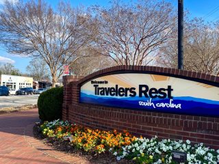 GVL A brick sign reads Welcome to Travelers Rest, South Carolina with flowers planted in front. Leafless trees and a few buildings, including the City Council offices, are visible in the background under a blue sky.
