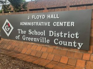 GVL A large sign in front of a brick wall reads: J. Floyd Hall Administrative Center, The School District of Greenville County, home to the Greenville County school board, with a logo featuring a G and a red flame.