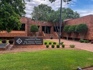 GVL A brick building with large windows and trees out front, home to the J. Floyd Hall Administrative Center. The sign displays “The School District of Greenville County”—where recent county funding cuts are a topic for the Greenville school board.