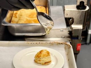 GVL A close-up of a hand holding a spoon, drizzling honey onto a biscuit on a white plate. A tray of more biscuits is visible in the background on a metal counter—a classic scene reminiscent of spoon bread origins.