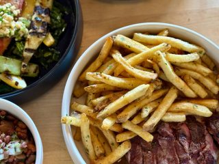 GVL A bowl filled with golden French fries and sliced steak sits beside a salad of grilled vegetables and a dish of beans on a wooden table, perfect for enjoying the Indaco supper series at The Commons.
