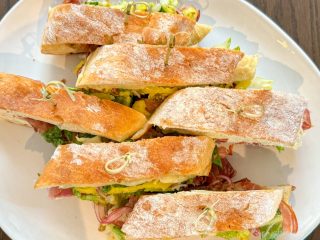 GVL Six rectangular sandwich halves with crusty, flour-dusted bread, filled with lettuce, cheese, and deli meat, are arranged on a white plate—an inviting choice from Tavola’s lunch menu.
