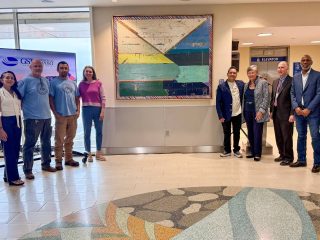 GVL A group of eight people stand in a semicircle around a large, colorful artwork by an Upstate artist at Greenville-Spartanburg airport, in a bright indoor space with a patterned floor and signage visible in the background.