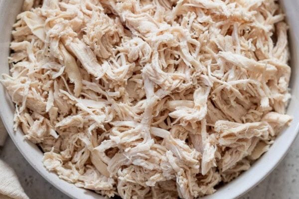 GVL A white bowl filled with Instant Pot shredded chicken sits on a light gray countertop next to a beige and brown striped cloth.