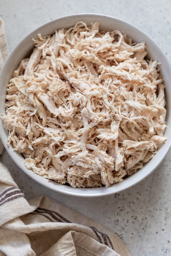 GVL A white bowl filled with Instant Pot shredded chicken sits on a light gray countertop next to a beige and brown striped cloth.