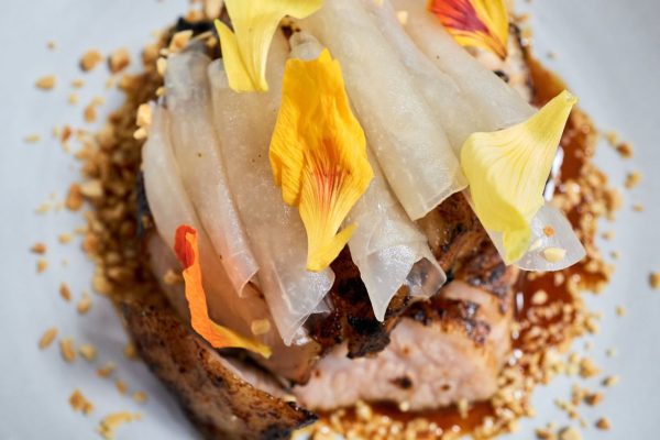 GVL A close-up of a gourmet dish at a dressed-up Taiwanese restaurant in Midtown, featuring seared fish topped with thin white vegetable slices, yellow and orange flower petals, chopped nuts, and a brown sauce on a white plate.