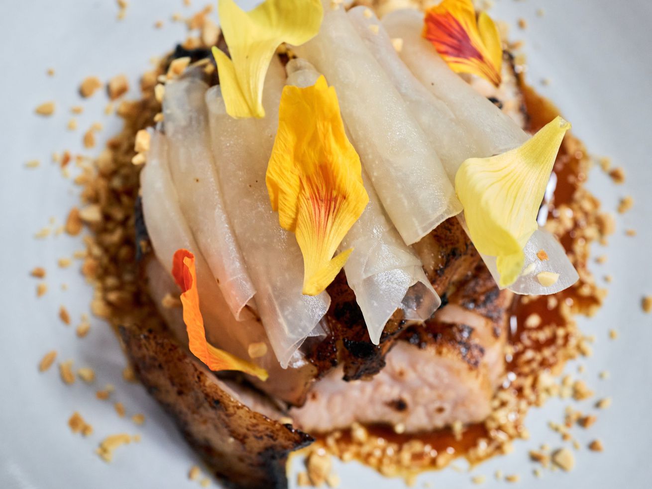 GVL A close-up of a gourmet dish at a dressed-up Taiwanese restaurant in Midtown, featuring seared fish topped with thin white vegetable slices, yellow and orange flower petals, chopped nuts, and a brown sauce on a white plate.