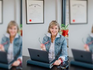 GVL A woman with short blonde hair, possibly Joan Herlong, sits at a desk with a laptop, holding eyeglasses and smiling. She wears a denim jacket; behind her are a framed certificate and window with potted plants—she’d never sell a house unprepared.