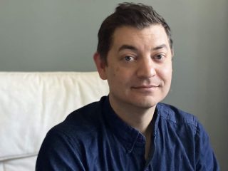 GVL A man with short brown hair wearing a dark blue button-up shirt sits on a white couch, smiling slightly, as if discussing Kevin Wilson’s new novel, with a neutral gray wall in the background.