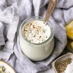 GVL A jar of creamy Blue Cheese Dressing topped with cracked black pepper and blue cheese crumbles, with a spoon inside. The jar sits on a striped cloth, surrounded by lemon wedges and small dishes of salt and pepper.