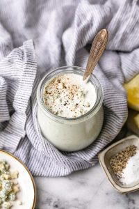 GVL A jar of creamy Blue Cheese Dressing topped with cracked black pepper and blue cheese crumbles, with a spoon inside. The jar sits on a striped cloth, surrounded by lemon wedges and small dishes of salt and pepper.