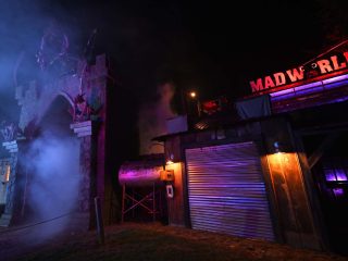 GVL A spooky, dimly lit entrance to Madworld haunted attraction with fog, a large arched gateway on the left, and a building with a glowing red MADWORLD sign on the right, celebrating 15 years of thrills.