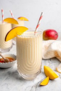 GVL A creamy mango smoothie in a clear glass with a striped straw and a mango slice on the rim. Fresh mango pieces and another glass of smoothie are in the background on a marble surface.