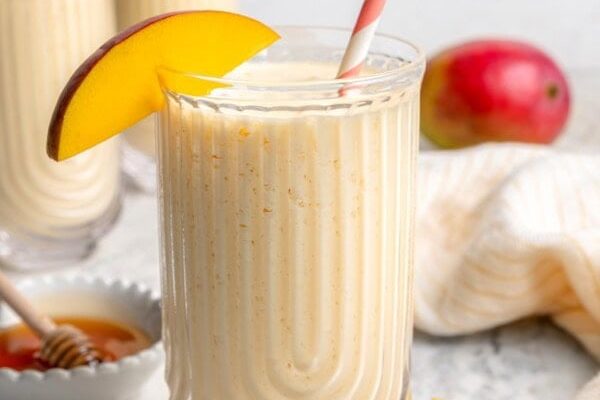 GVL A creamy mango smoothie in a clear glass with a striped straw and a mango slice on the rim. Fresh mango pieces and another glass of smoothie are in the background on a marble surface.