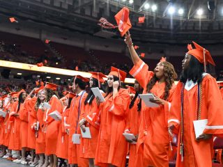 GVL Mauldin graduates in orange caps and gowns stand in a row at an indoor graduation ceremony. Some toss their caps in the air, celebrating the Class of 2025, while others smile and hold diplomas under stadium lights.