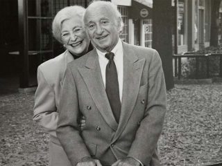 GVL Max and Trude Heller stand outdoors, smiling warmly at the camera. The man wears a suit and tie, while the woman stands behind him in a light jacket. Fallen leaves surround them, reflecting their lasting Greenville legacy.