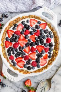 GVL A No-Bake Yogurt Pie with a date-nut crust features a creamy white filling, topped with sliced strawberries, blueberries, blackberries, and raspberries. It rests on a marble surface beside a striped cloth and serving utensil.
