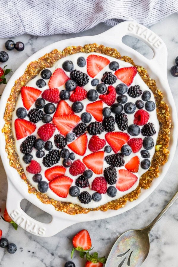 GVL A No-Bake Yogurt Pie with a date-nut crust features a creamy white filling, topped with sliced strawberries, blueberries, blackberries, and raspberries. It rests on a marble surface beside a striped cloth and serving utensil.
