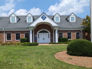 GVL A brick building with white columns and a blue and white City Hall logo above the entrance, surrounded by green grass, bushes, and a partly cloudy sky—an example of Simpsonville development.