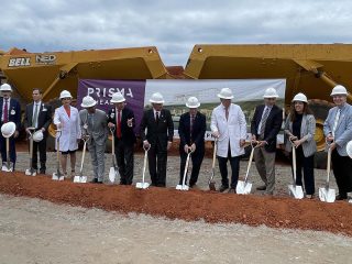 GVL A group of people in business attire and hard hats stand in a line with shovels at the groundbreaking ceremony for Prisma’s $138M behavioral health hospital, with large construction vehicles and a Prisma Health banner in the background.