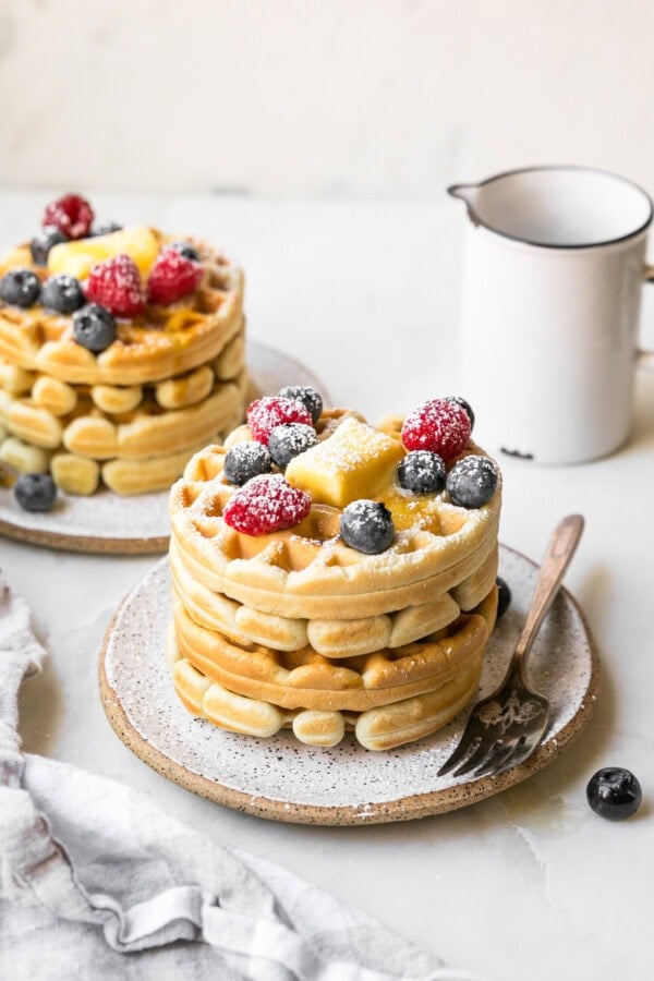 GVL Two stacks of golden sourdough waffles topped with mixed berries, butter, and powdered sugar sit on ceramic plates. A fork rests on one plate, and a white pitcher is in the background on a white surface.