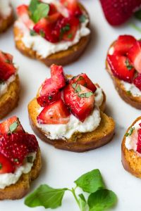 GVL Toasted bread slices topped with creamy ricotta, fresh diced strawberries, and chopped basil make this irresistible Strawberry Crostini—a perfect strawberry appetizer for any gathering. Arranged on a white surface with a sprig of green herbs nearby.