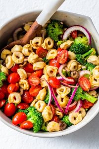 GVL A vibrant Tortellini Pasta Salad with cherry tomatoes, broccoli, red onion slices, and olives is mixed together in a bowl with a wooden spoon, all arranged on a clean white surface.