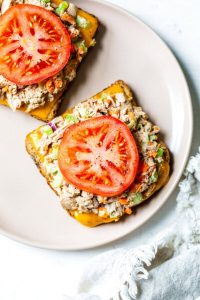 GVL Two open-faced tuna melts on a beige plate, each topped with a slice of tomato, tuna salad, and melted cheese on toasted bread. A white textured cloth is partially visible beside the plate.